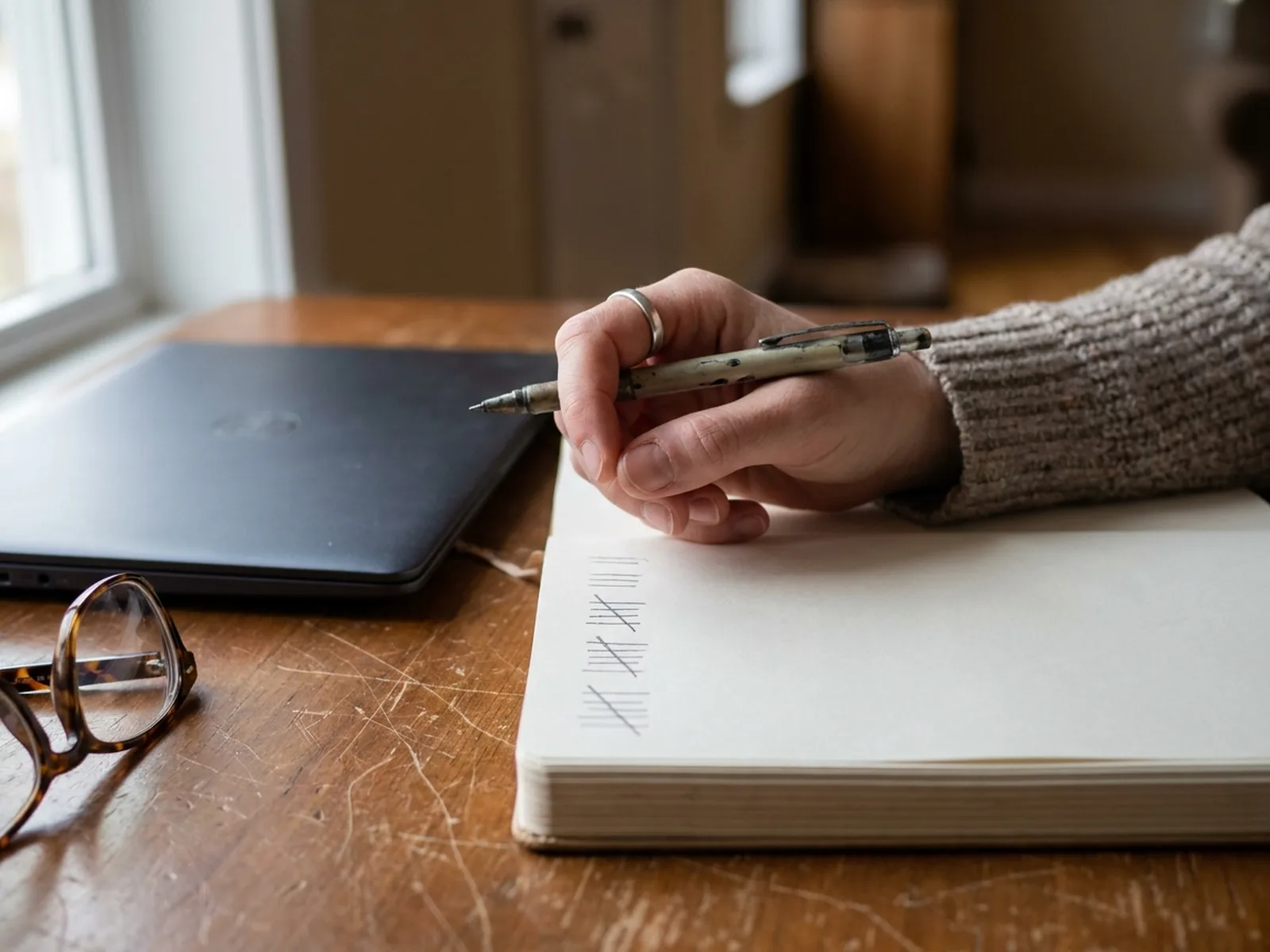 A hand keeping a tally in a notebook beside a laptop and a pair of glasses
